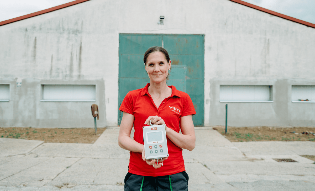 A lady standing infront of a poultry house holding a BAT1 manual poultry scale in her hands